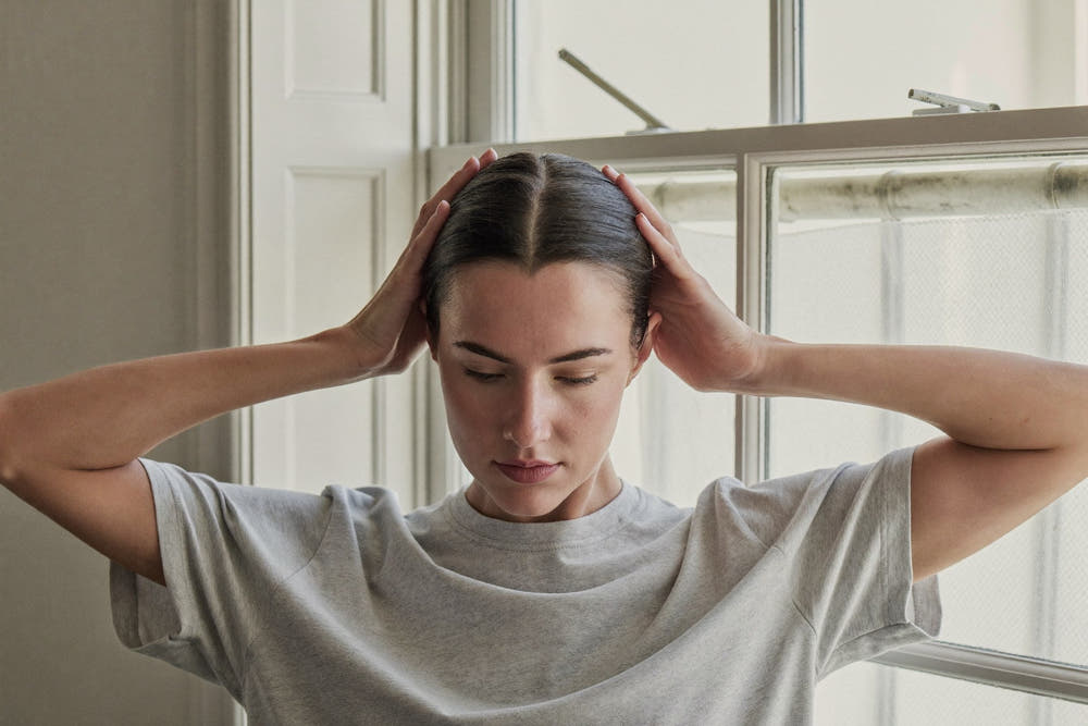 A woman in a soft grey t-shirt with both hands at her parted dark hair, fingertips on the scalp, by a bright window