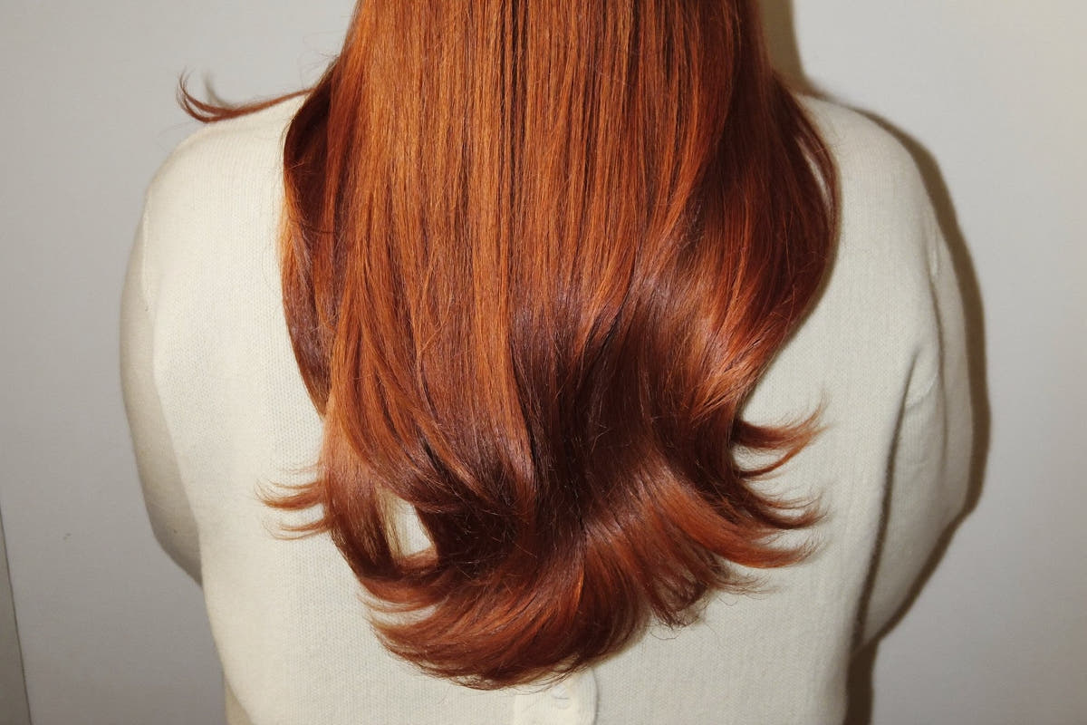 Long glossy copper-red dyed hair seen from behind, on a woman in a cream knit cardigan, in soft light