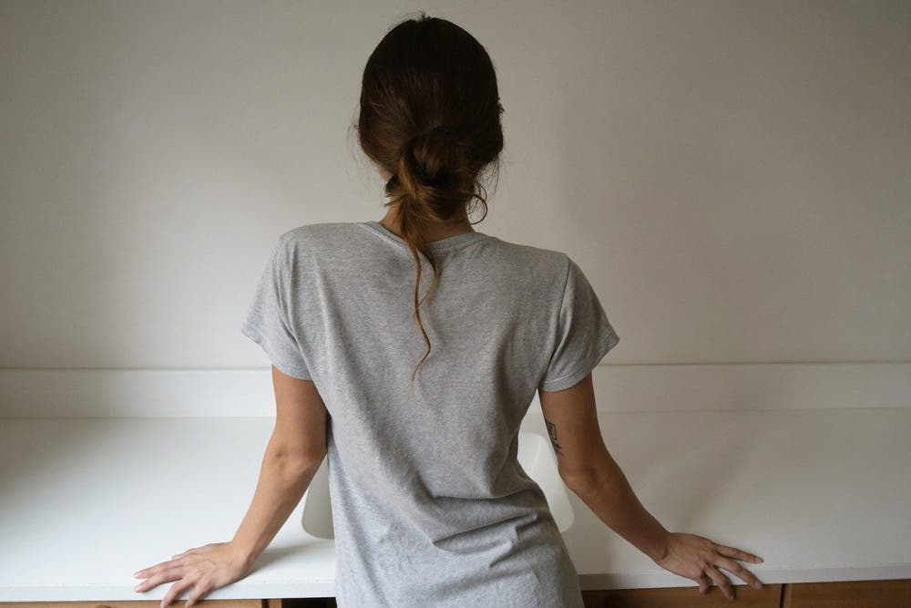 A woman in a soft grey t-shirt, seen from behind, leaning on a white bathroom sink in morning light, her hair in a loose bun