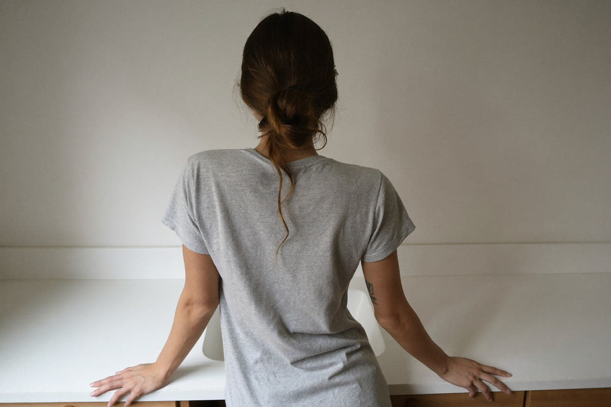 A woman in a soft grey t-shirt, seen from behind, leaning on a white bathroom sink in morning light, her hair in a loose bun