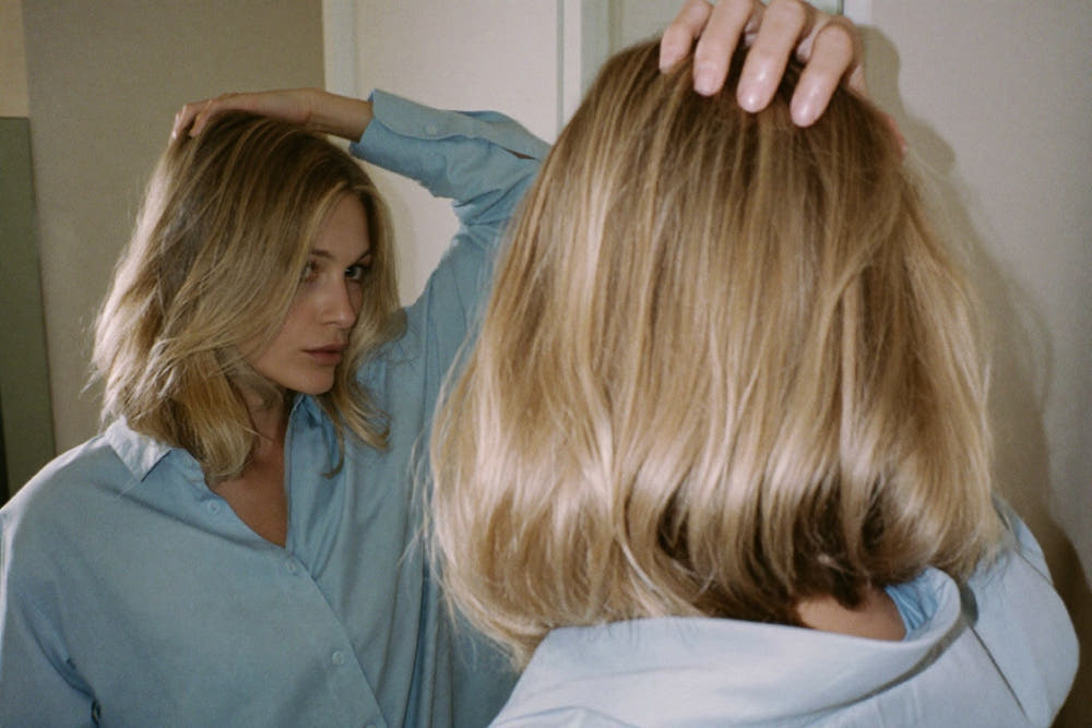 A woman with fine, straight blonde hair lifting her roots with her fingers in front of a mirror