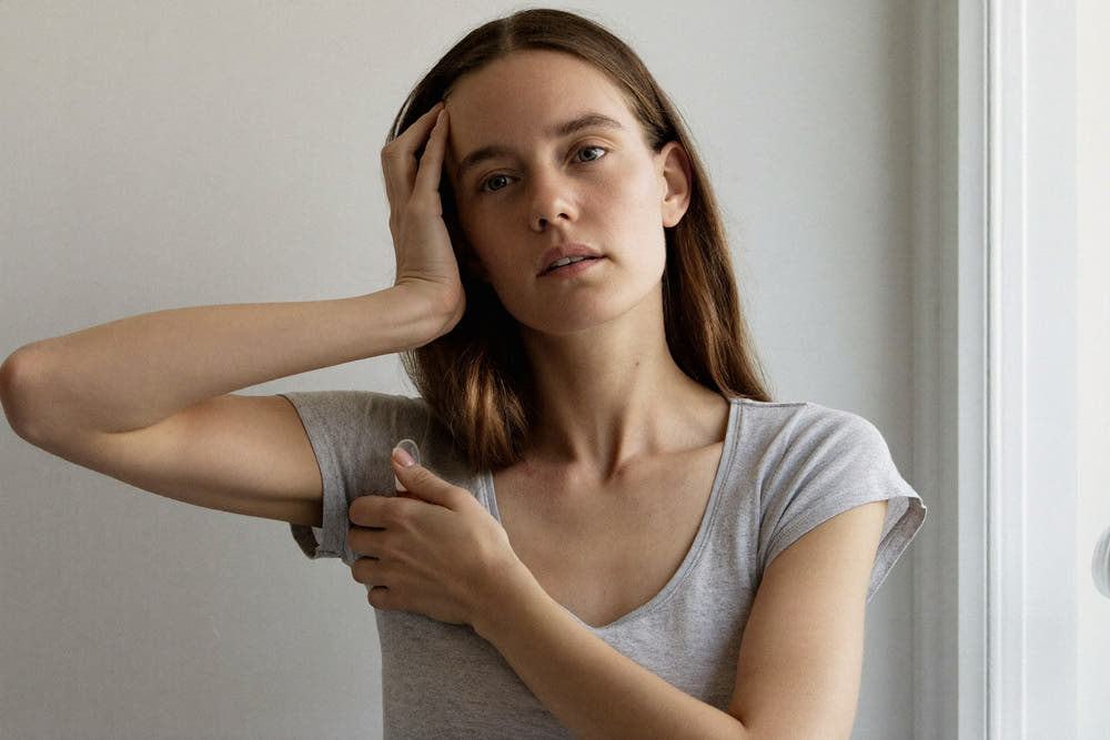 A woman in a soft grey t-shirt touching her hairline with her fingertips in early morning light