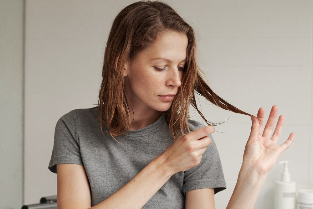 A woman with damp chestnut hair smoothing the ends between her fingers in a bright bathroom