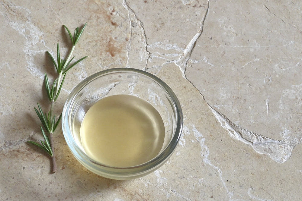 A shallow glass dish of pale liquid beside a fresh rosemary sprig on a pale stone surface in soft daylight
