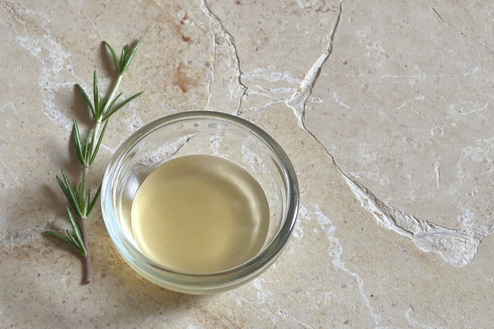 A shallow glass dish of pale liquid beside a fresh rosemary sprig on a pale stone surface in soft daylight