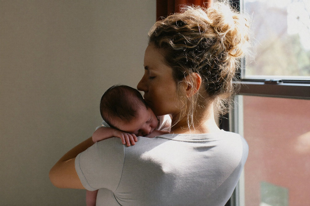 A woman in a soft grey t-shirt holding a sleeping baby on her shoulder by a bright window, her hair in a loose bun