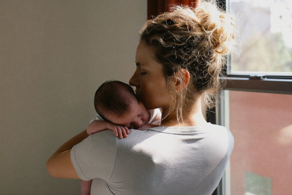 A woman in a soft grey t-shirt holding a sleeping baby on her shoulder by a bright window, her hair in a loose bun