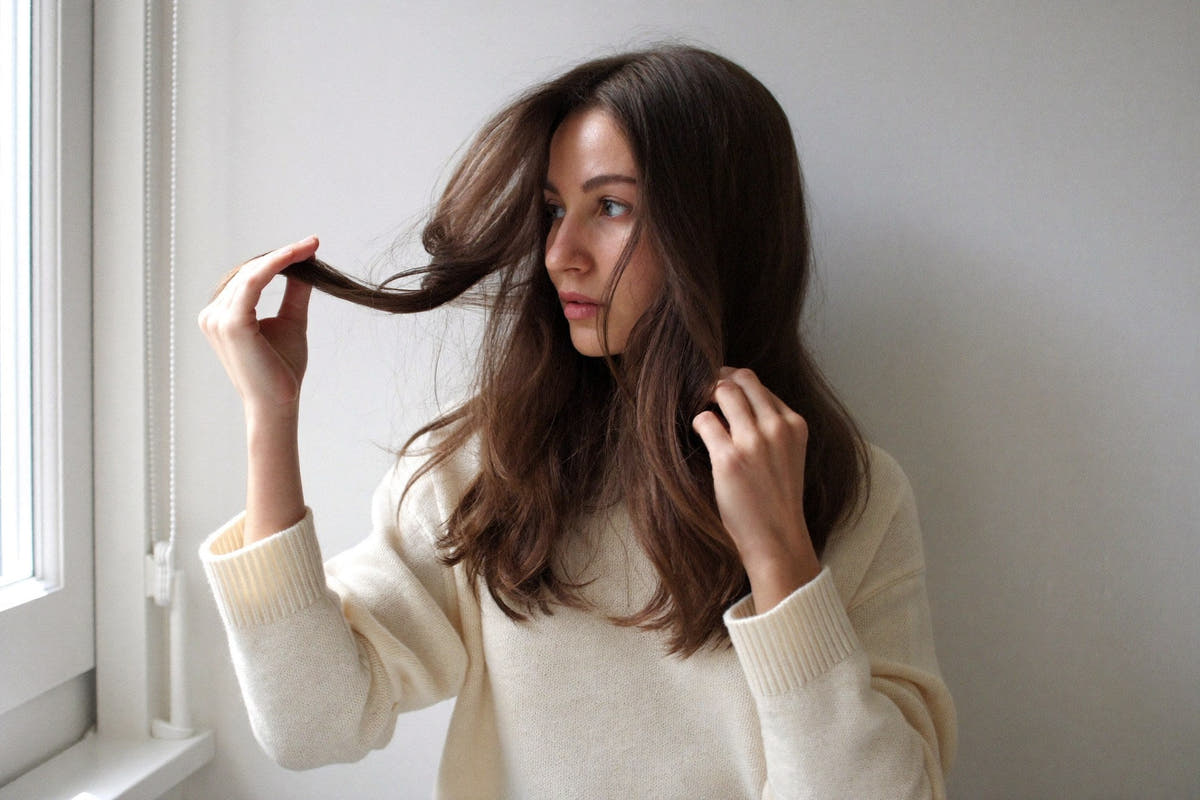 A woman in a cream knit sweater holding the ends of her long hair up to the light, looking at them