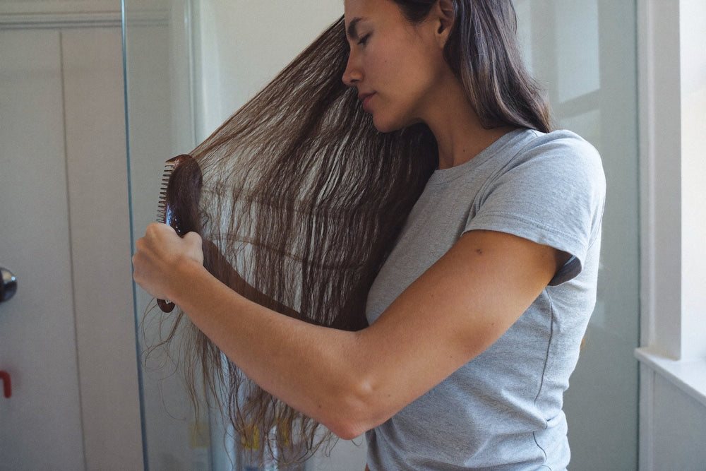A woman in a grey t-shirt combing through long dry brown hair in a bright bathroom