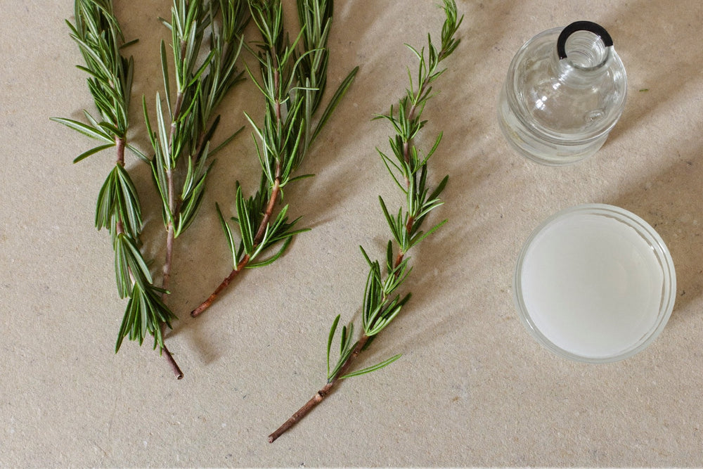 Fresh rosemary sprigs on a pale stone surface beside a small glass jar and a dish of pale liquid