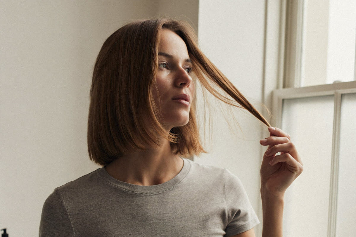A woman with fine brown hair by a window, holding a strand out to examine it