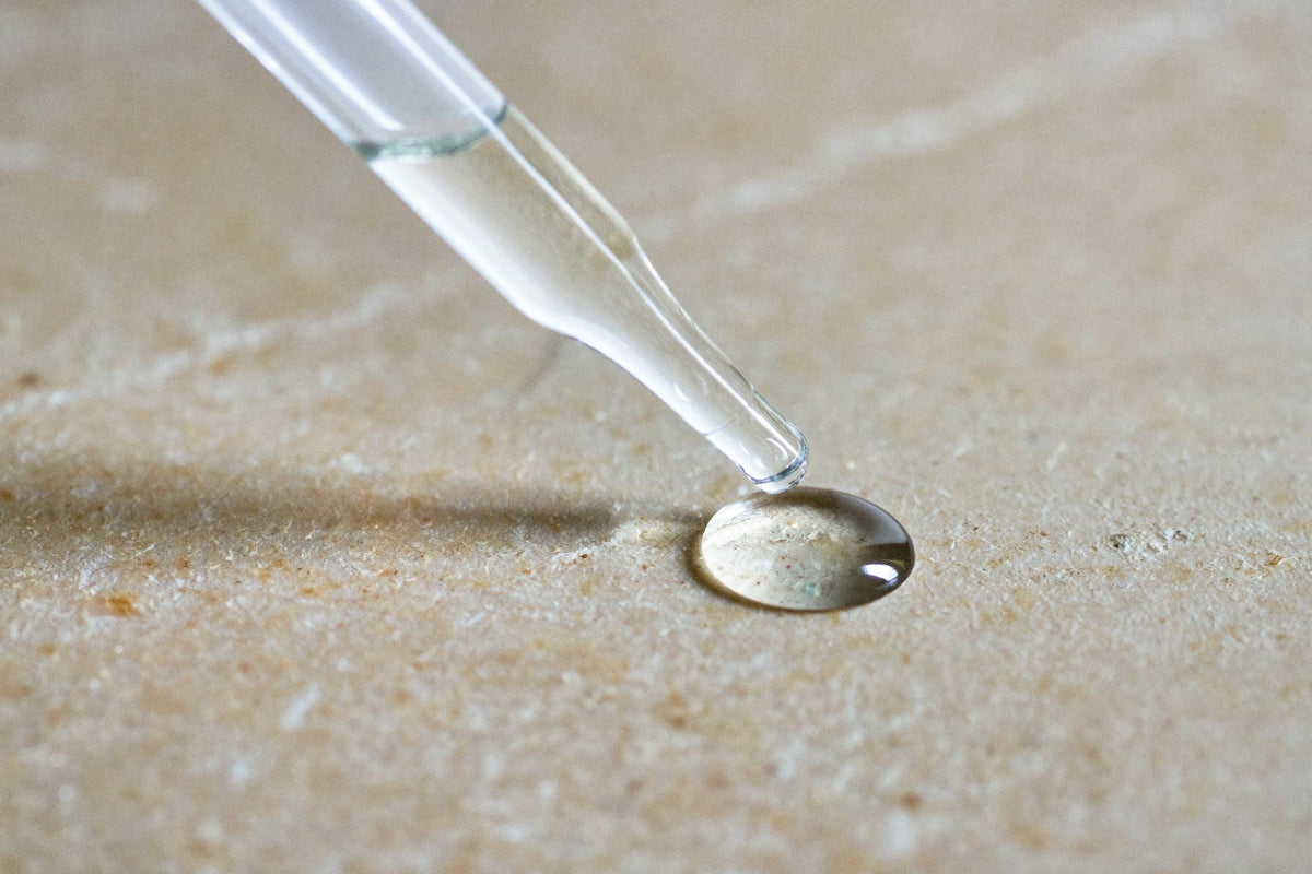 A glass pipette releasing a single clear drop onto a pale stone surface