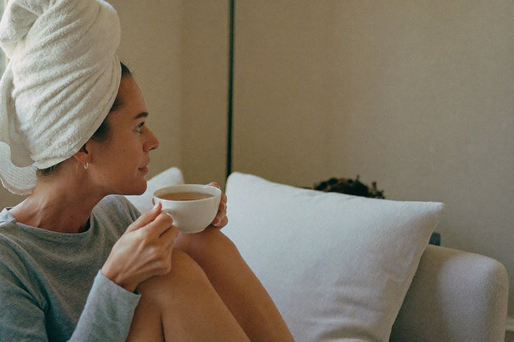 A woman with her hair wrapped in a towel, sitting on a sofa with a cup of tea