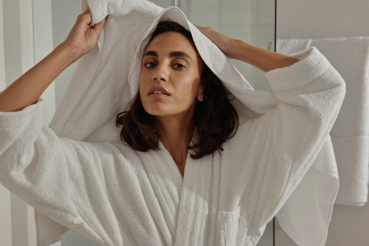 A woman in a white cotton robe towel-drying her hair in a bright, slightly steamy bathroom