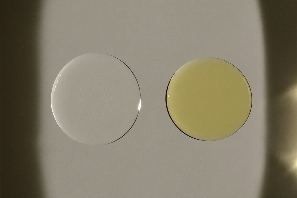 Two drops on a pane of glass, lit from the side: one a clear round bead, one a golden oil drop spreading thin